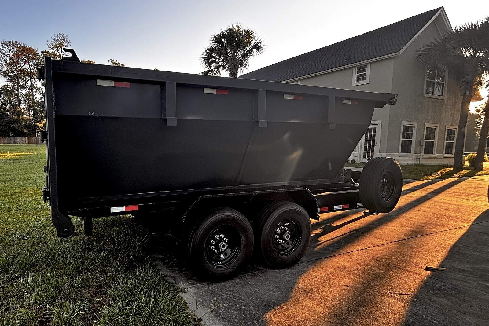 Black roll-off container with high steel sides, delivered and parked on a driveway at sunset.
